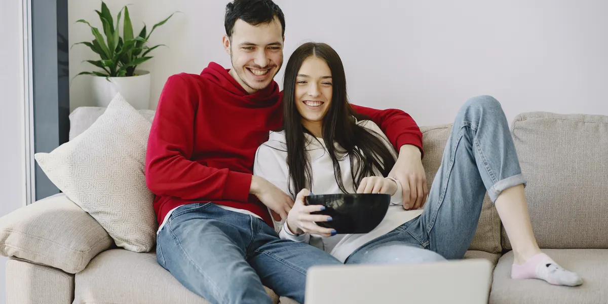 A young couple laughing together on a pale sofa while watching a laptop, a snack bowl held between them